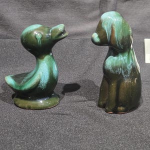 May include: Two ceramic figurines: a duck and a dog. The duck has a long neck and open beak, while the dog sits upright. Both are glazed in shades of green and teal, with a glossy finish. The duck is approximately 3 inches tall.