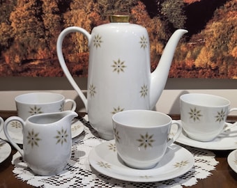 May include: A white porcelain tea set with a gold-trimmed coffee pot, creamer, cups, and saucers. Each piece is decorated with a repeating floral pattern in gold and olive green. The set is displayed on a white lace tablecloth.