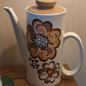 Puede incluir: Cafetera de cerámica color crema con tapa y asa marrones. La cafetera está decorada con grandes diseños florales estilizados en marrón y naranja. El pico es largo y delgado.