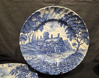 May include: Blue and white decorative plate featuring a detailed illustration of Anne Hathaway's Cottage. The plate has a scalloped edge and a classic, vintage aesthetic. The scene includes trees, a cottage, and figures.