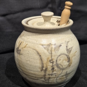 Może przedstawiać: Beżowy ceramiczny słoik z pokrywką i małą drewnianą łyżką. Słoik ma okrągły kształt z lekko teksturowaną powierzchnią i jest ozdobiony brązowymi i beżowymi abstrakcyjnymi wzorami. Pokrywka ma mały uchwyt.