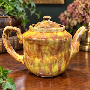 May include: A ceramic teapot featuring a glossy, mottled glaze in shades of vibrant yellow, deep orange, and earthy brown. The teapot has a rounded body, a curved handle, a short spout, and a matching lid with a small knob on top.