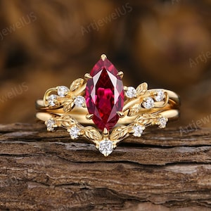Peut inclure: Une bague en or avec une pierre précieuse rouge foncé taille marquise au centre. L'anneau et l'alliance en forme de V assortie sont ornés de petites feuilles en or sculptées et de minuscules pierres blanches rondes sur les branches.