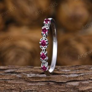 May include: A silver-toned ring featuring a series of four round-cut, vibrant pinkish-red gemstones set in a row. The metal band displays intricate, interconnected knot-like metalwork detailing between each of the circular gemstone settings.