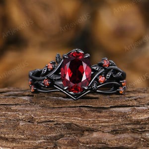 Könnte beinhalten: Ein schwarzer Ring mit einem großen, oval geschliffenen roten Edelstein. Der Ring hat ein florales Design mit kleineren roten Akzentsteinen und liegt auf einer strukturierten Holzoberfläche.