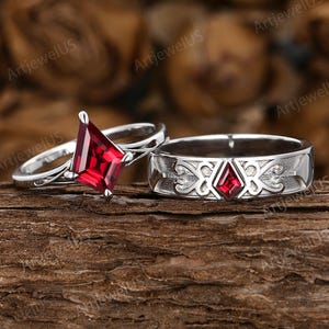May include: Two silver rings with red gemstones. One ring features a kite-shaped gemstone, while the other has a smaller, square-cut stone set in an engraved band. The rings are displayed on a wooden surface, with the text "ArtjewelUS" visible.