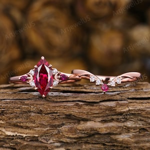 May include: A rose gold ring set featuring a marquise-cut red centre stone flanked by smaller red and clear stones, paired with a curved band accented with clear stones and a single red stone, all set against a textured wood surface.