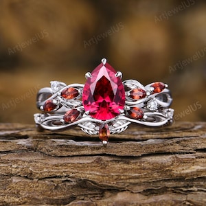 Peut inclure: Une bague argentée ornée d'une grande pierre rouge taille poire au centre. L'anneau présente un travail de métal complexe en forme de feuilles, agrémenté de plusieurs petites pierres rouges taille marquise et de minuscules pierres rondes claires.