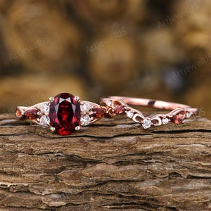 May include: A rose gold ring set featuring a central oval-cut deep red garnet flanked by smaller marquise-cut garnets and round white stones. The matching curved band includes intricate metalwork with embedded round and marquise-cut stones.