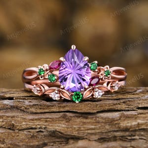 May include: A rose gold ring set featuring a central pear-cut purple amethyst, accented with small round green emeralds, marquise-cut red rubies, and clear white diamonds arranged in a floral-inspired band design with intricate metal leaf details.