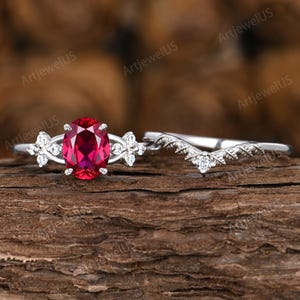 Op de afbeelding: Een tweedelige ringenset met een zilveren band met een grote ovaal geslepen robijn edelsteen en een bijpassende gebogen band met kleine accentstenen. De ringen zijn geplaatst tegen een getextureerde houten achtergrond.