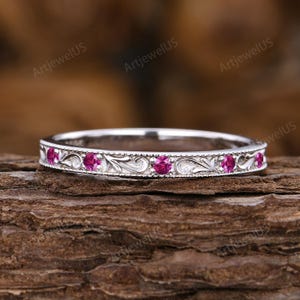 Peut inclure: Une bague en argent ornée d'une série de petites pierres précieuses roses rondes. La bande est décorée d'un motif gravé et orné. La bague est présentée sur une surface en bois brun texturé.