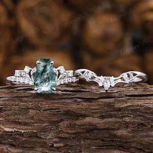 May include: A two-piece ring set featuring a silver band with a rectangular, green-hued gemstone and a matching curved band. The main ring has a central stone with a unique pattern, surrounded by smaller, clear stones. The curved band is adorned with similar clear stones.