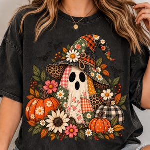 Peut inclure: Un t-shirt gris anthracite orné d'un motif représentant un fantôme portant un chapeau de sorcière en patchwork. Le fantôme est entouré de citrouilles orange et à carreaux noirs et blancs, de tournesols, de marguerites, de fleurs roses et de chauves-souris.