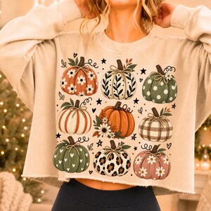 Peut inclure: Un sweat-shirt court beige à manches longues avec un imprimé graphique de neuf citrouilles aux motifs variés, incluant des fleurs, des pois, des carreaux et des taches de léopard, agrémenté de petites étoiles et cœurs noirs.