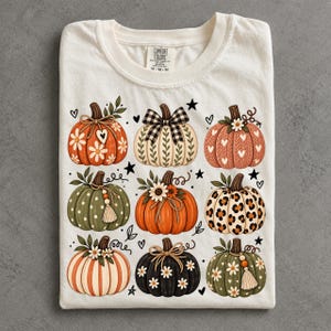 Peut inclure: Un t-shirt à col rond blanc cassé avec une impression graphique de neuf citrouilles stylisées disposées en grille. Les citrouilles présentent divers motifs dont des marguerites, des imprimés léopard, des nœuds vichy, des cœurs et des pompons.