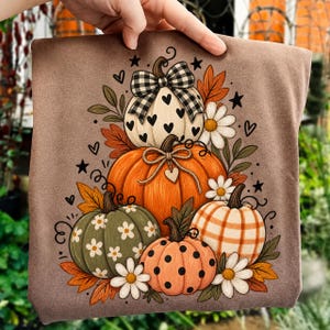 Peut inclure: Un t-shirt marron avec une illustration graphique d'une pile de cinq citrouilles. Les citrouilles sont décorées de motifs à carreaux noirs et blancs, de cœurs blancs, de marguerites, de carreaux orange et de pois noirs, avec des feuilles d'automne.