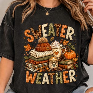 Peut inclure: Un t-shirt noir avec une impression graphique portant le texte "SWEATER WEATHER". Le motif comprend une pile de pulls pliés, un bonnet en tricot, une tasse de chocolat chaud avec de la crème fouettée, une citrouille, une bougie et des feuilles d'automne.