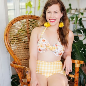 Könnte beinhalten: Eine Frau in einem gelben und weißen Gingham-Bikini-Oberteil mit Blumendruck und passenden Höschen sitzt auf einem Korbstuhl. Sie trägt gelbe Quastenohrringe und roten Lippenstift.