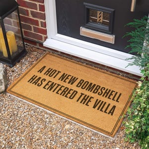 May include: A rectangular tan coir doormat with a black border and the black text A HOT NEW BOMBSHELL HAS ENTERED THE VILLA printed in the center. The mat has a coarse, fibrous texture and is positioned horizontally on a gravel surface.