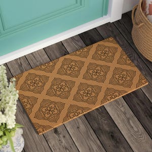 May include: A rectangular coir doormat featuring a repeating pattern of dark brown, stylized floral motifs set against a natural tan background. The design consists of symmetrical, square-shaped ornaments with central flower shapes and intricate borders.