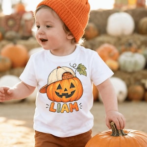 May include: A white short-sleeved t-shirt featuring a graphic of a smiling jack-o'-lantern wearing a beige baseball cap, with the name LIAM printed in colorful block letters underneath the pumpkin illustration.