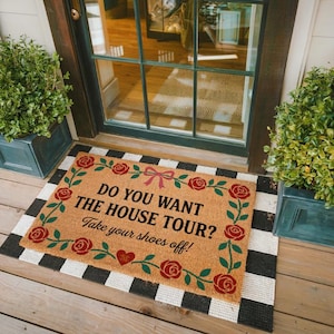 May include: A rectangular doormat with the text "DO YOU WANT THE HOUSE TOUR? Take your shoes off!" in black. The mat is tan with a border of red roses and green leaves. The border is framed by a black and white checkered pattern.