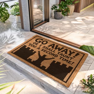 May include: A rectangular tan coir doormat featuring a dark brown silhouette of a castle and a figure with raised arms. The text reads: GO AWAY! OR I SHALL TAUNT YOU A SECOND TIME! The mat has a dark brown border and is placed on a tiled floor.