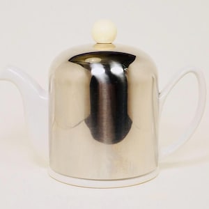 May include: A silver-colored teapot with a white base, handle, and spout. The lid has a small, round, cream-colored knob. The teapot has a reflective surface, and the handle is curved.