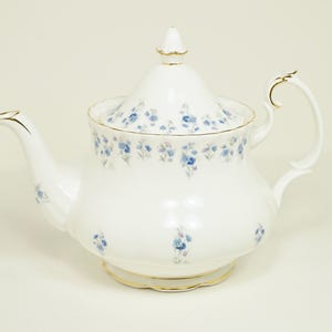 May include: White porcelain teapot with a lid, decorated with blue floral patterns. The teapot has a curved spout, a handle, and gold trim. A small gold finial sits atop the lid. A classic tea accessory.
