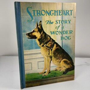 Può includere: Copertina di un libro vintage con un cane pastore tedesco con un'imbracatura. Il titolo recita "Strongheart The Story of a Wonder Dog" in lettere dorate e turchesi. Il libro ha un dorso blu ed è ambientato su uno sfondo azzurro e verde.
