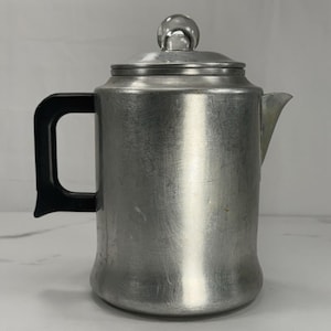May include: A brushed aluminum coffee percolator featuring a cylindrical body, a black heat-resistant handle, and a matching lid topped with a rounded knob. The metal surface shows light signs of wear and texture consistent with vintage kitchenware.