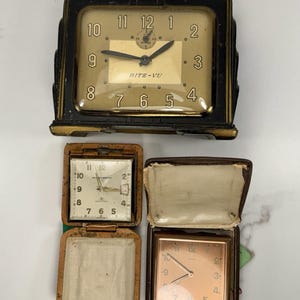 Op de afbeelding: Een verzameling vintage klokken. De bovenste klok is zwart en goudkleurig met de tekst "RITE-VU" op de wijzerplaat. Twee kleinere reiskokken zitten in leren etuis, één open en één gesloten. De klokken hebben verschillende gekleurde wijzerplaten.