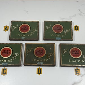 May include: Five vintage Lucky Strike cigarette tins. Each tin is rectangular with a green background and a red and gold circular logo. The text "LUCKY STRIKE" and "CIGARETTES" are visible. The word "flat" is written on each tin.