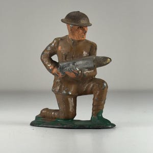 May include: A vintage toy soldier figurine in a kneeling position, painted in shades of brown and tan. The soldier wears a helmet and uniform, holding a silver-colored weapon. The base is green.