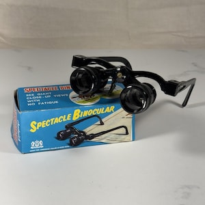 May include: Black spectacle binoculars with a blue box. The box reads "Spectacle Binocular" in yellow and white text. The binoculars are designed to provide giant close-up views with no fatigue.
