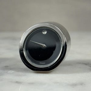 Könnte beinhalten: Eine Movado-Uhr mit schwarzem Zifferblatt und silbernem Gehäuse. Die Uhr hat ein minimalistisches Design mit einem einzelnen Punkt auf 12 Uhr und silbernen Zeigern. Das Wort "MOVADO" ist am unteren Rand des Zifferblatts aufgedruckt.