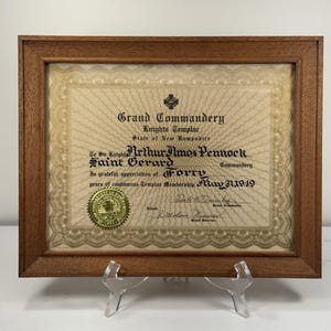 May include: A framed certificate from the Grand Commandery Knights Templar, State of New Hampshire. The certificate, dated May 31, 1949, is in a brown wooden frame and features ornate text and a gold seal. The certificate is propped up on a clear plastic stand.