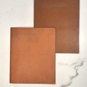 May include: Two brown, rectangular notebooks are stacked on a white marble surface. The notebooks have a worn appearance, with visible text on the covers. The color is a warm, earthy tone.