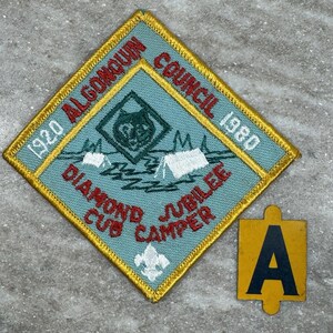 May include: A vintage Algonquin Council Cub Camper patch with a diamond shape, featuring a wolf emblem, tents, and text. The patch is bordered in gold and includes the text "1920 Algonquin Council 1980 Diamond Jubilee Cub Camper". A yellow "A" tag is next to the patch.