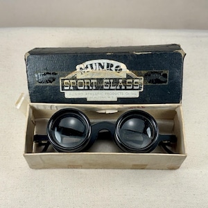 May include: Vintage black Munro Sport Glass binoculars in their original box. The box is black with white text and the binoculars are in a tan cardboard box. The binoculars have large, round lenses and a black frame.