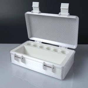 May include: A white rectangular storage case with a hinged lid and two top latches. The interior features a large open compartment and a row of six circular indentations along the back edge. The exterior surface has a textured, grid-like pattern.