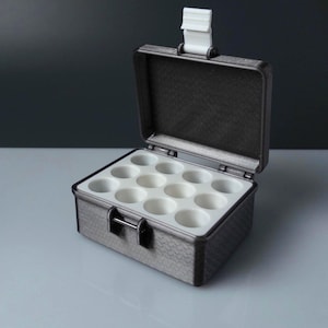 May include: A dark grey rectangular storage case with a textured exterior and a hinged lid. The interior features a white insert with twelve circular indentations arranged in a grid. A white latch is positioned at the top centre of the lid.