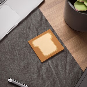 May include: A square sticky note with a tan bread slice design. The bread slice has a light tan center and a darker tan outline, set against a brown background. The note is on a dark gray surface.