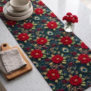 May include: A dark teal table runner with a floral pattern of large red roses, peacock feathers, and berries. The runner is on a light-colored countertop, with a stack of plates and a bowl, a wooden cutting board, and a vase of red flowers.