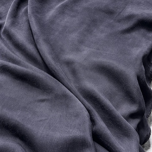 K&ouml;nnte beinhalten: Nahaufnahme eines drapierten, dunkelgrauen Stoffes mit seidiger Textur. Der Stoff weist weiche Falten und subtile Lichtreflexe auf, die ein Gef&uuml;hl von Tiefe und Bewegung erzeugen. Das Material wirkt leicht und flie&szlig;end.