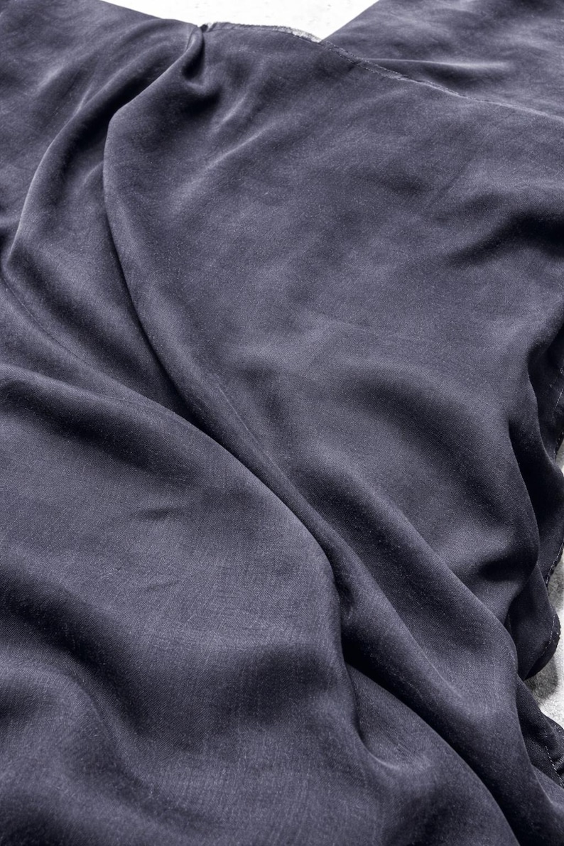 K&ouml;nnte beinhalten: Nahaufnahme eines drapierten, dunkelgrauen Stoffes mit seidiger Textur. Der Stoff weist weiche Falten und subtile Lichtreflexe auf, die ein Gef&uuml;hl von Tiefe und Bewegung erzeugen. Das Material wirkt leicht und flie&szlig;end.