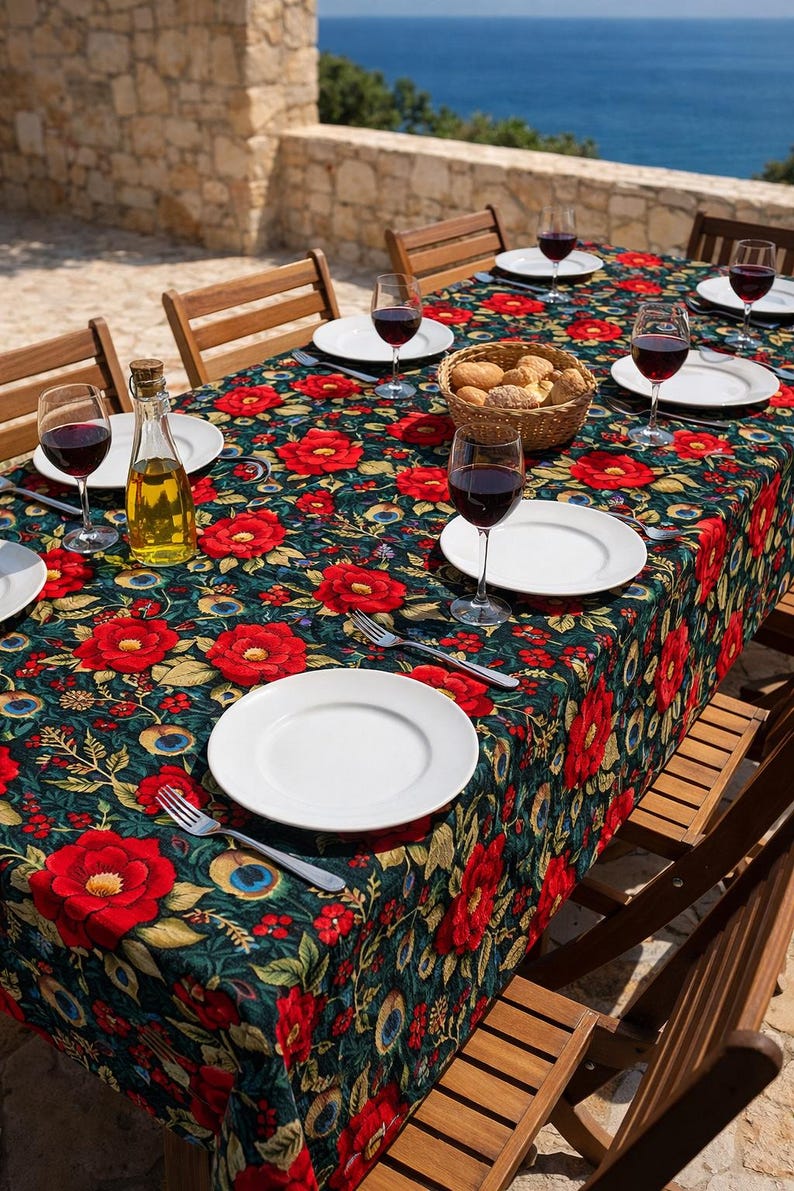 May include: A dining table set for a meal outdoors, featuring a floral tablecloth with red flowers and green leaves. White plates, wine glasses with red wine, and a basket of bread are arranged on the table. Wooden chairs surround the table.