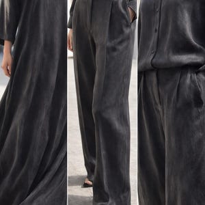 K&ouml;nnte beinhalten: Ein dunkelgraues Ensemble mit einem flie&szlig;enden V-Ausschnitt-Kleid, einer hoch taillierten Hose mit Falten und einem Hemd mit Kn&ouml;pfen. Die Kleidung scheint aus einem weichen, flie&szlig;enden Stoff zu bestehen, der f&uuml;r eine minimalistische Garderobe geeignet ist.