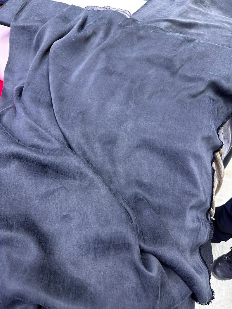 K&ouml;nnte beinhalten: Nahaufnahme eines gro&szlig;en St&uuml;cks dunkelblauen Stoffes, m&ouml;glicherweise Seide oder ein &auml;hnliches Material. Der Stoff ist drapiert und gefaltet, mit sichtbarer Textur und subtilen Farbvariationen. Die Kanten des Stoffes sind sichtbar und zeigen die rohen, unfertigen Kanten.